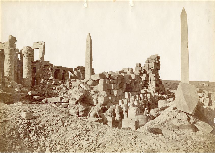 The photograph shows a view from the south of the Wadjet Hall of the Karnak Temple Complex, built by Pharaoh Tuthmosis I, as well as the two obelisks of Hatshepsut (one standing, the other collapsed). In the background in the centre, the surviving obelisk of Tuthmosis I is also visible. An egyptian sits on one of the plinths of the massive columns. The author's signature is visible at the bottom right.