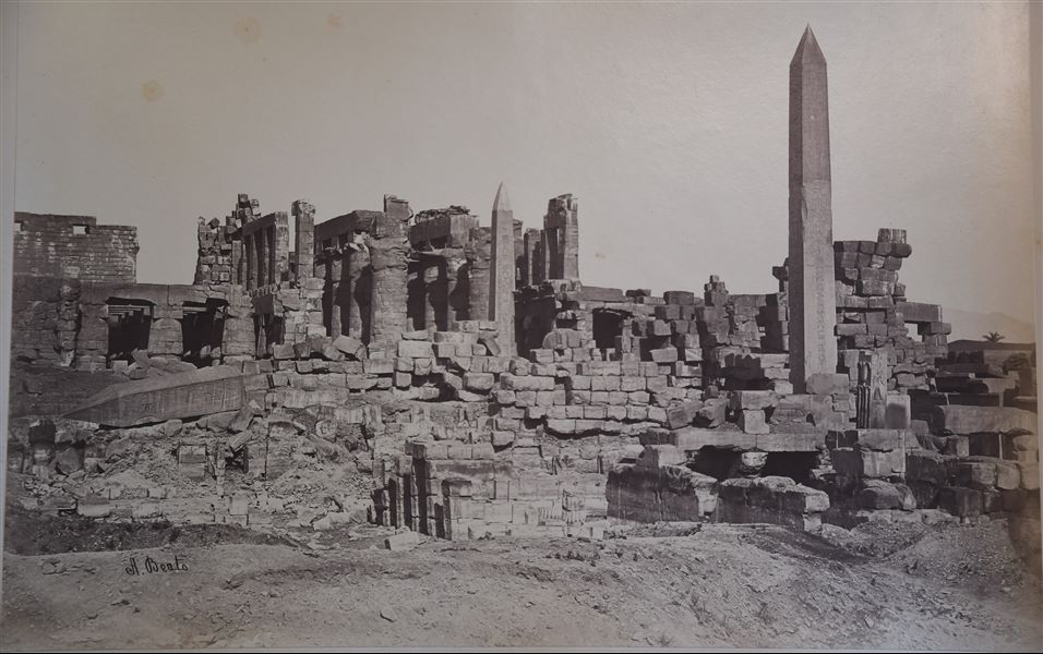 Veduta dei resti della Grande Sala Ipostila, davanti alla quale sono ben visibili i tre obelischi, due ancora eretti (uno di Thutmosi I, al centro, l’altro di Hatshepsut, a destra) e uno crollato (anch’esso della regina Hatshepsut), in seguito ad un terremoto nel Medioevo. La firma dell’autore si trova in basso a sinistra.
