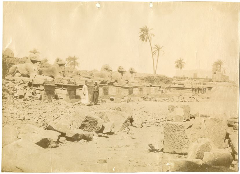 The photograph shows the ram-headed sphinxes of the great avenue leading to the first pylon of the Karnak Temple Complex, with two local inhabitants among the ruins of the ancient monuments. The photo can be attributed to Antonio Beato.   