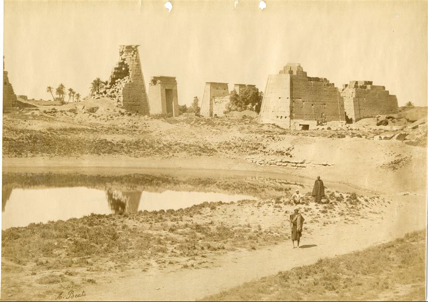 La fotografia ritrae le rovine di due piloni del complesso templare di Amon a Karnak, visti dal Lago Sacro, in particolare il IX pilone (a sinistra, del faraone Horemheb) e l'VIII pilone (a destra, della regina Hatshepsut). Al centro, sullo sfondo, sono visibili la porta meridionale voluta da Tolemeo III Evergete per l’accesso al tempio di Khonsu, del quale si vede il pilone: la prospettiva è falsata, ma i due monumenti sono allineati. Tre egiziani in riva al Lago Sacro, lievemente prosciugato, guardano verso l'obiettivo. In basso a sinistra è riconoscibile la firma dell’autore. 
