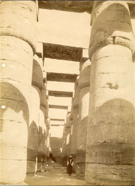 View of the colonnade of the Great Hypostyle Hall of the Karnak Temple Complex, where a local inhabitant poses in its shadow. Note that the ground level still covers the columns’ bases. The author's signature can be found at the bottom centre.   