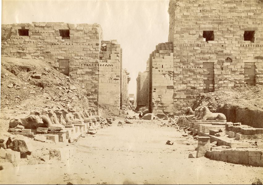 The image depicts the façade of the first pylon of the Karnak Temple Complex, photographed from the Avenue of the Sphinxes, with a figure sitting among the ruins. In the background, the central corridor of the Great Hypostyle Hall can be seen, aligned with the entrance to the temple. The photograph can be attributed to Antonio Beato.   