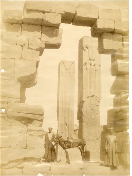 The image shows the ruins of the vestibule built by Tuthmosis III in the Karnak Temple Complex. In particular, the monolithic pink granite pillars bearing the emblems of Upper Egypt (right) and Lower Egypt (left) stand out, with the Lotus and Papyrus respectively. The photo can be attributed to Antonio Beato. 