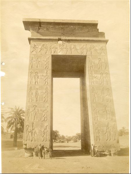 A photograph of the western gate to the Karnak Temple Complex, built by Pharaoh Ptolemy IV. Some Egyptians and a donkey are posing in front of it, giving an idea of the colossal proportions. The author's signature is visible at the bottom left. 