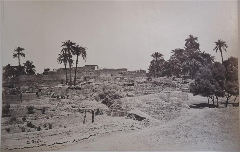 Veduta paesaggistica del villaggio di Karnak, a Luxor. Sullo sfondo si intravedono i resti del complesso templare di Amon, tra cui spicca un obelisco (a sinistra), e la struttura imponente della grande sala ipostila. La firma dell’autore si trova in basso a sinistra.