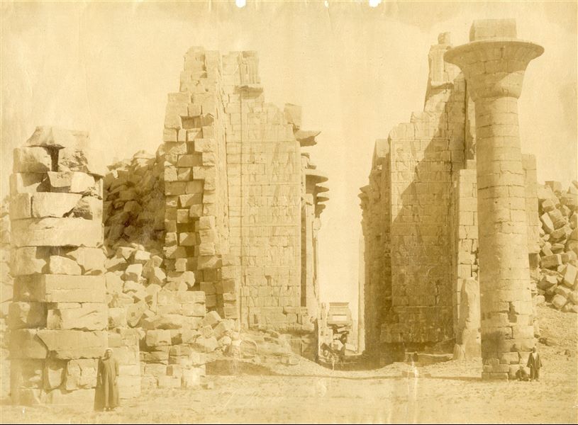 The picture shows the entrance to the Great Hypostyle Hall of the Karnak Temple Complex, seen from the first courtyard. The remaining column from the south-eastern corner of the kiosk of the 25th dynasty Pharaoh Taharqa stands out. Some locals give an idea of the colossal size of the complex. The author's signature is visible at the bottom.