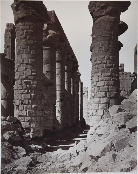 Detail of the central nave of the Great Hypostyle Hall from the Karnak Temple Complex, in the direction of the second pylon. The ground level still partially covers the columns' bases. The author's signature can be found at the bottom right. 