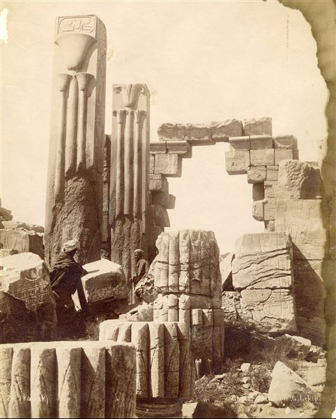The image shows the ruins of the vestibule built by Tuthmosis III in the Karnak Temple Complex, where the pink granite pillars bearing the emblems of Upper Egypt (right) and Lower Egypt (left) are visible, respectively, the lotus for the South and the papyrus for the North. Above, the cartouche of Tuthmosis III. Two egyptians are present at the foot of the columns. The authors' signature is legible at the bottom right. 