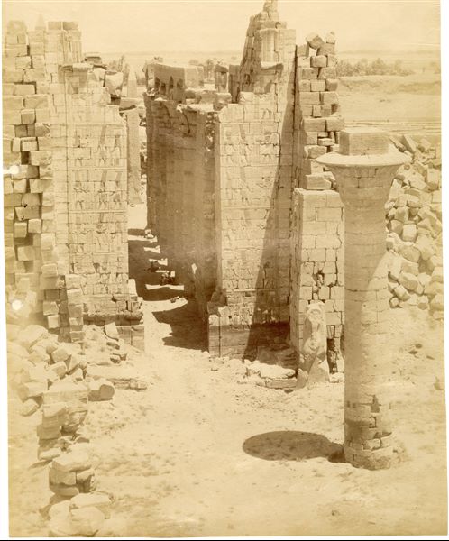 Il primo cortile del complesso templare di Amon a Karnak è qui fotografato dal I pilone, attraverso il quale si accede al tempio. Si possono riconoscere la colonna rimasta del chiosco del faraone Taharqa (XXV dinastia, detta “etiope”) e il corridoio centrale della Grande Sala Ipostila, costruita dal faraone Seti I. Sullo sfondo si riconoscono i due obelischi, ancora presenti: quello del faraone Tutmosi I (al centro) e quello della regina Hatshepsut (di cui si vede solo la cuspide, a sinistra). La firma dell'autore, benché molto schiarita e quasi illeggibile, è posta in basso a destra.
