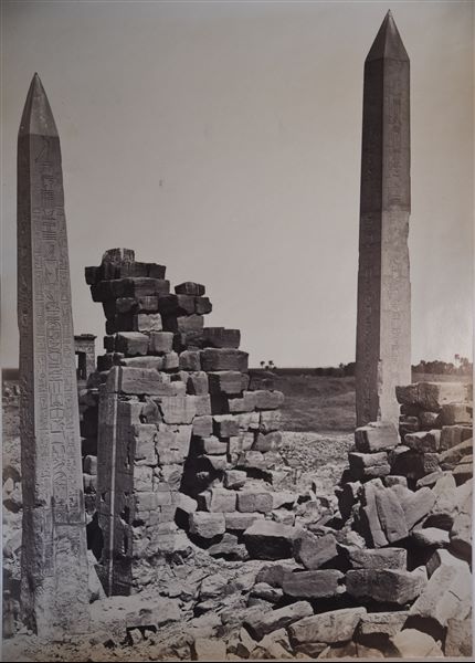 Veduta dei due obelischi ancora in posizione eretta nel complesso templare di Amon a Karnak, uno del faraone Thutmosi I (a sinistra), l’altro della regina Hatshepsut (a destra), in mezzo si trovano invece i resti del IV pilone. In basso a destra, la firma dell’autore.  