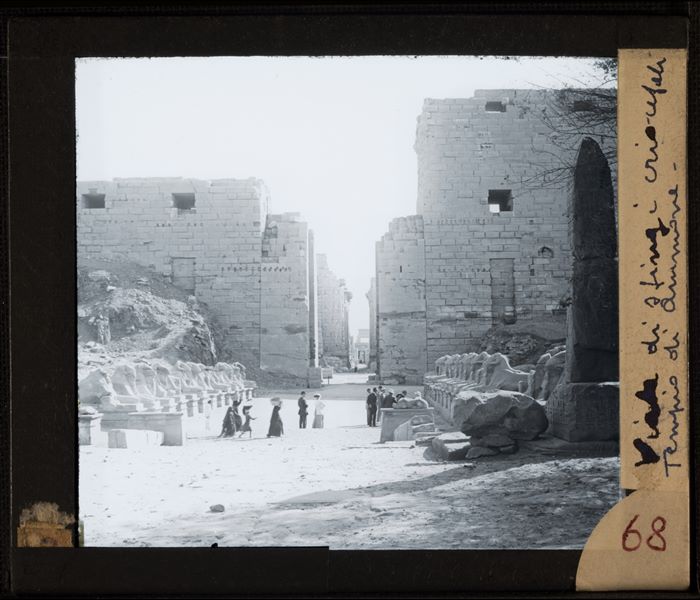Western entrance to the Karnak Temple Complex. In the foreground on the left, a pylon and on either side of the road a series of ram-headed sphinxes. In the avenue, there is a group of Egyptians at work (left) and a group of Europeans visiting the ruins on the right.