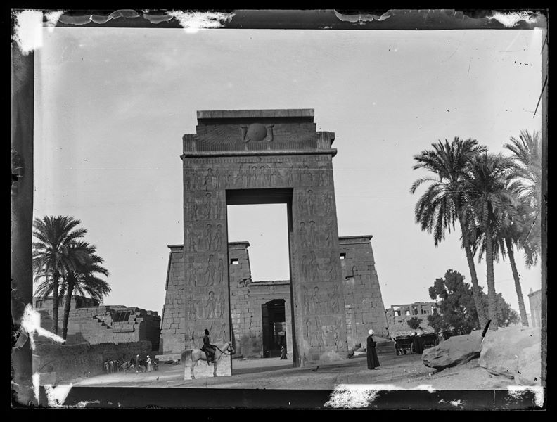  Karnak, image showing the entrance gateway of Ptolemy III Euergetes. In the foreground, there is one Egyptian man standing and another on horseback, most likely a soldier.
