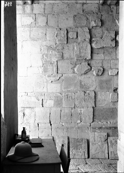 South side of the mortuary temple of Queen Hatshepsut at Deir el-Bahari. Wall scene from the portico in the middle terrace depicting a village in the region of Punt. In the foreground, a small table with some stationery materials and a colonial hat.