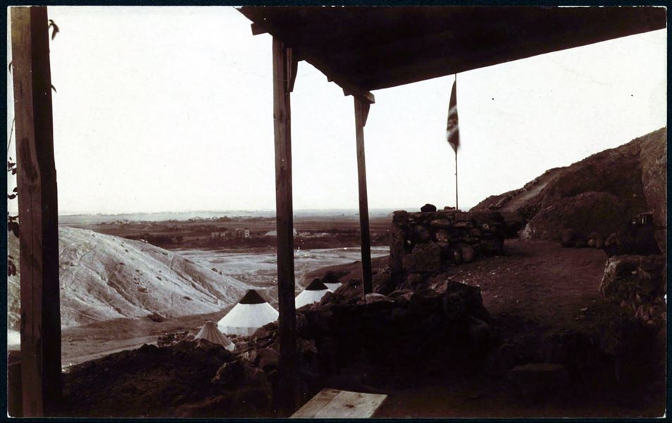 Veranda della casupola in mattoni crudi della Missione Archeologica Italiana durante gli scavi a Deir el-Medina, nel 1909. Su un livello inferiore, si contano quattro tende coniche. Sullo sfondo è visibile la piana verdeggiante della riva ovest del Nilo, e i resti architettonici del tempio di Medinet Habu, costruito da Ramesse III. Scavi Schiaparelli 