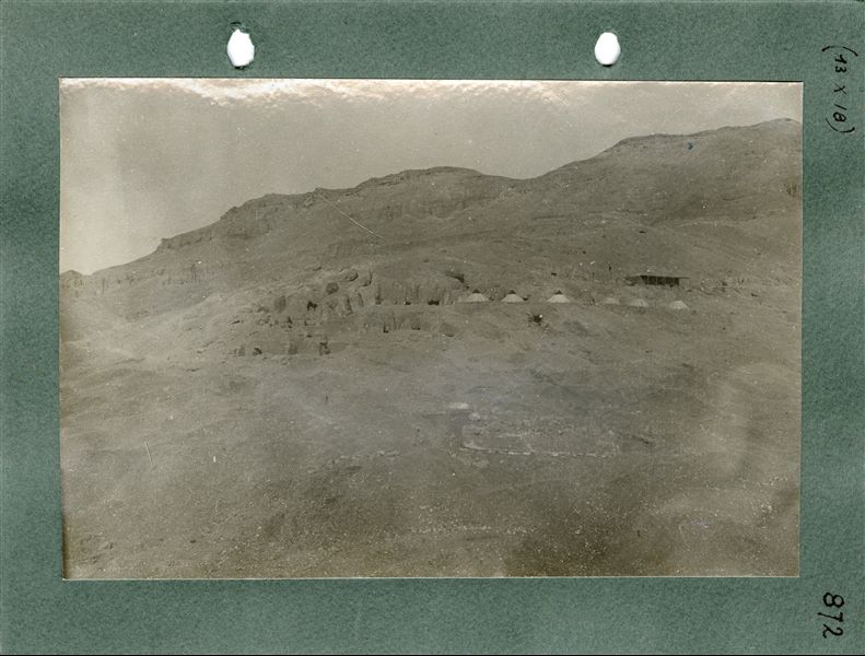 Fotografia del villaggio di Deir el-Medina, appena visibile in primo piano, e veduta dell’accampamento della missione italiana, composto da sei tende coniche, e da una piccola costruzione in mattoni con veranda, su un livello superiore del declivio. Sullo sfondo, la montagna di Deir el-Medina, dove furono scavate nella roccia numerose tombe. Scavi Schiaparelli 
