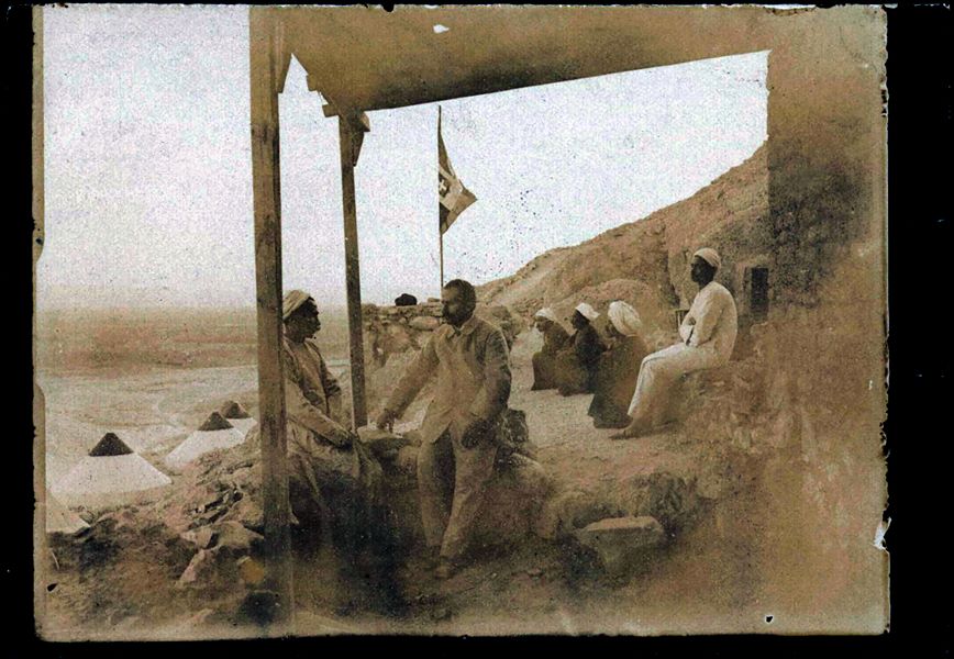 Veranda della piccola costruzione in mattoni crudi della Missione Archeologica Italiana durante gli scavi a Deir el-Medina, nel 1909. Nella fotografia sono immortalati alcuni membri della missione archeologica, non riconosciuti. Il personaggio al centro non è al momento riconoscibile, mentre il giovane a destra potrebbe essere l’aiuto cuoco, Buhus. Su un livello inferiore, si contano quattro tende coniche. Sullo sfondo è visibile la piana verdeggiante della riva ovest del Nilo. Fotografia poco nitida. Scavi Schiaparelli 