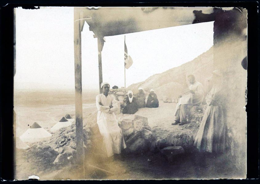 Veranda della piccola costruzione in mattoni crudi della Missione Archeologica Italiana durante gli scavi a Deir el-Medina, nel 1909. Nella fotografia sono presenti alcuni membri della missione archeologica, non riconosciuti. Il personaggio seduto mentre legge il giornale non è al momento riconoscibile, mentre il giovane a sinistra potrebbe essere l’aiuto cuoco, Buhus. Su un livello inferiore, si contano quattro tende coniche. Sullo sfondo è visibile la piana verdeggiante della riva ovest del Nilo. Fotografia poco nitida. Scavi Schiaparelli 