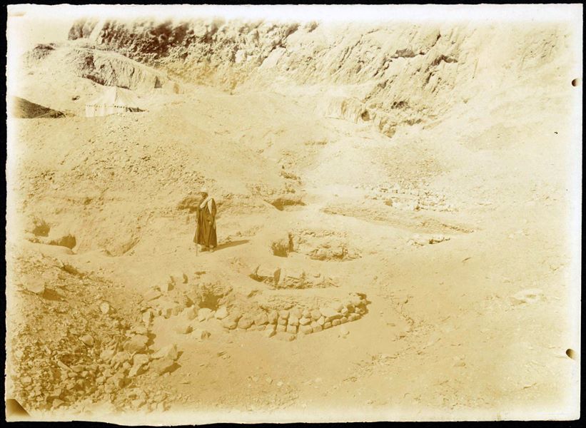 Photograph taken during excavations to the north of the temple of Hathor, where its shadow can be seen on the left. One of the tents used by the Italian Archaeological Mission is also visible on the left. The figure in the centre is unidentifiable. Photograph slightly overexposed. This print on paper has been toned with sepia. Schiaparelli excavations.