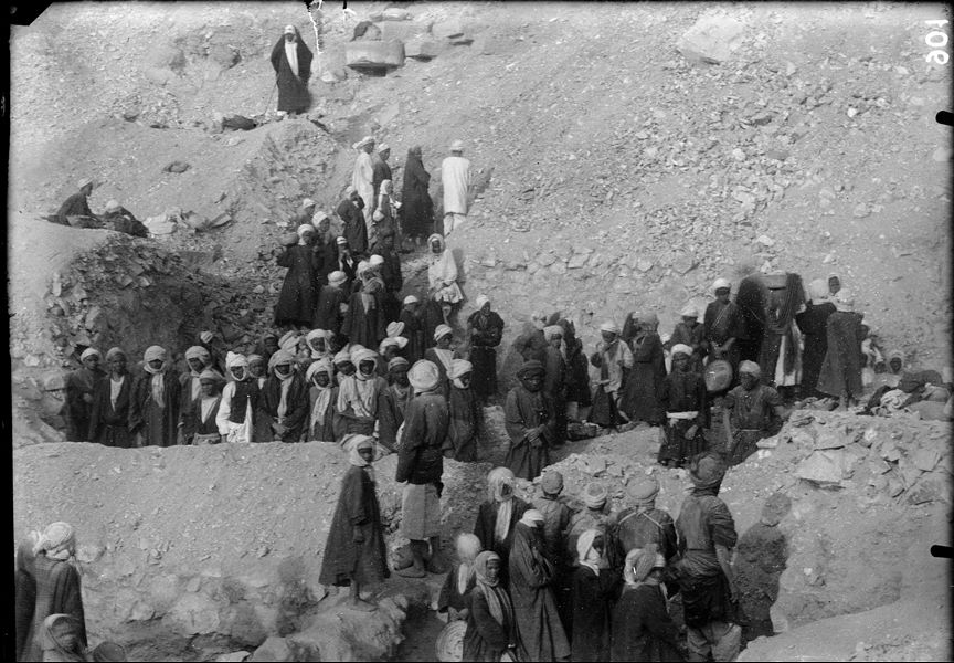 Excavations along the mountain, behind the temple of the goddess Hathor. The workmen have stopped excavating in order to photograph the site without dust-clouds. Above, the figure of the rais (chief of the workmen). Schiaparelli excavations. 