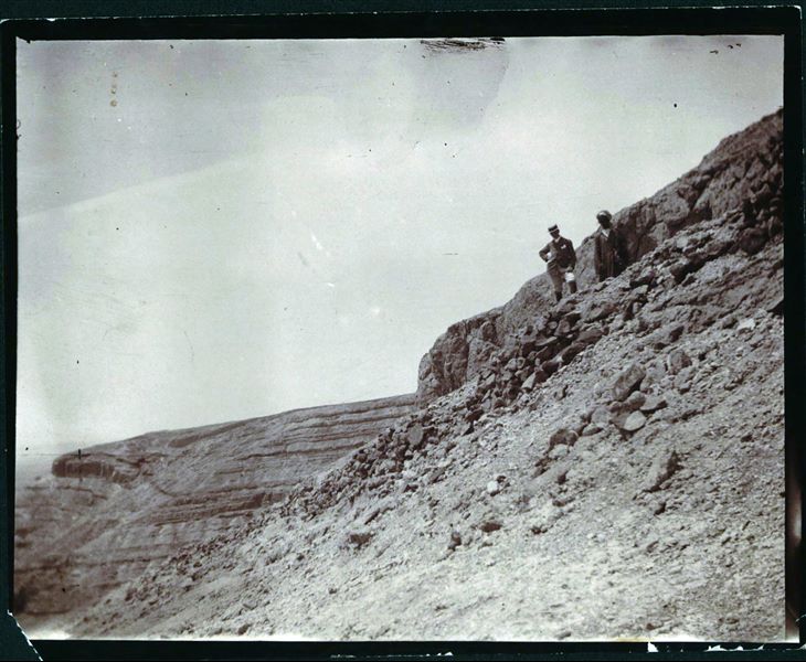 Declivio di una montagna, sulla quale sono presenti due personaggi, non identificati ma presumibilmente parte della Missione Archeologica Italiana. Il personaggio con abiti occidentali potrebbe essere uno dei nobili al seguito di Schiaparelli nella missione del 1906. Scavi Schiaparelli.