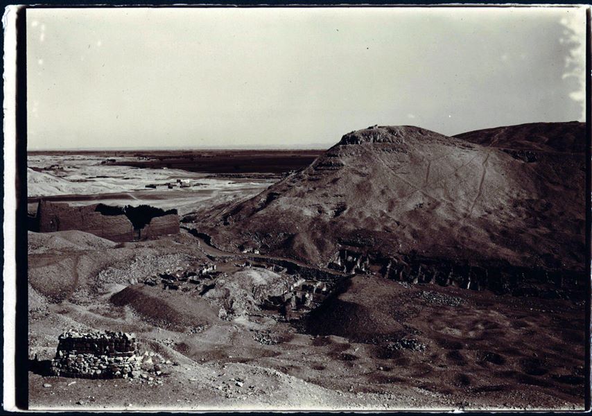 Veduta generale dell’area di Deir el-Medina. La foto è stata scattata dalla montagna a occidente, in basso si vedono le prime case del villaggio con a sinistra, il tempio della dea Hathor. Di fronte la montagna orientale che delimita il sito, sulla sinistra si intravedono spuntare le rovine del Ramesseo con i suoi magazzini. Sullo sfondo infine la pianura coltivata fino a raggiungere il Nilo. Fotografia di poco diversa (leggermente a sinistra) rispetto a C02043. Scavi Schiaparelli.