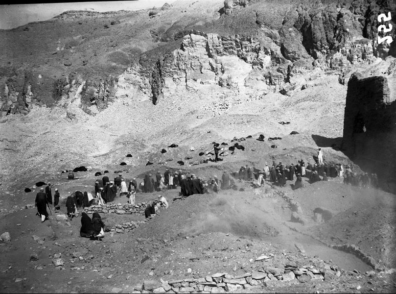 Excavations near the left boundary wall of the temple of the goddess Hathor. Workers are excavating among the remains of ancient buildings. In the centre of the photo a member of the mission is surveying the site. On the left there is a large vase (ziro), containing water for the workers. Schiaparelli excavations. 