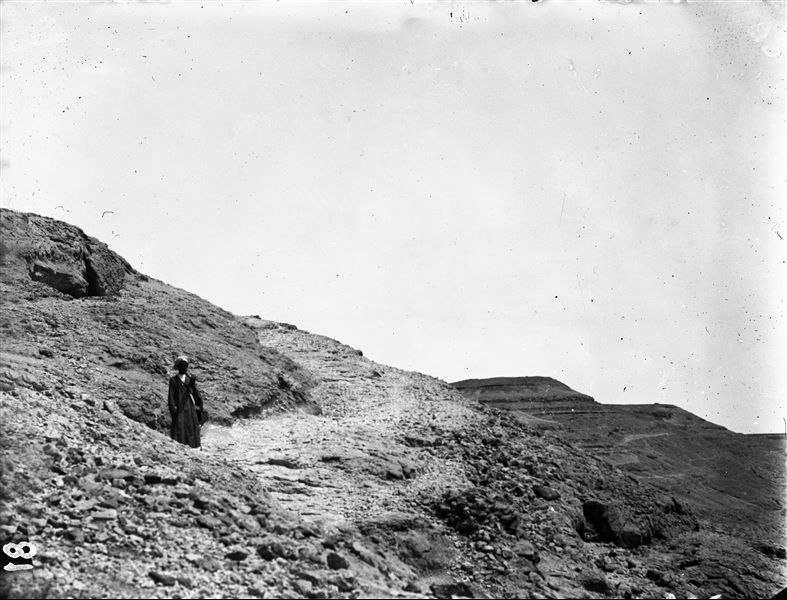 Montagna a settentrione dell’area di Deir el-Medina. L’immagine riprende un camminamento che conduce alla sommità della montagna. L’uomo fotografato porta a tracolla un tubo per disegni o carte geografiche e tre macchine fotografiche di cui una automatica a rullino. Scavi Schiaparelli. 