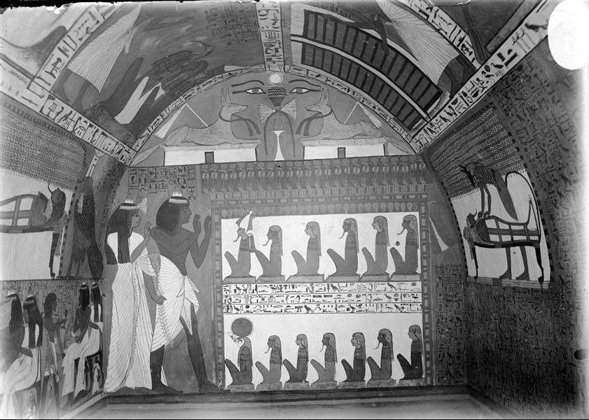 West wall (left) from the tomb of Sennedjem (TT1). Above, two jackals seated on a shrines; the lower register shows the deceased  Sennedjem together with his wife Iyneferti as they worship the gods of the Duat, including Osiris (the first in the top row from the left) and Re-Horakhty (the first in the bottom row from the left). 