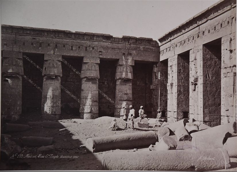 View of the second court of the Temple of Medinet Habu, built by Pharaoh Ramesses III on the west bank of the Nile at Thebes. Several columns can still be seen on the ground, with the standing papyrus columns still partially buried, which help to date the photo to before the site was cleaned and restored. The author's signature is visible at the bottom. 