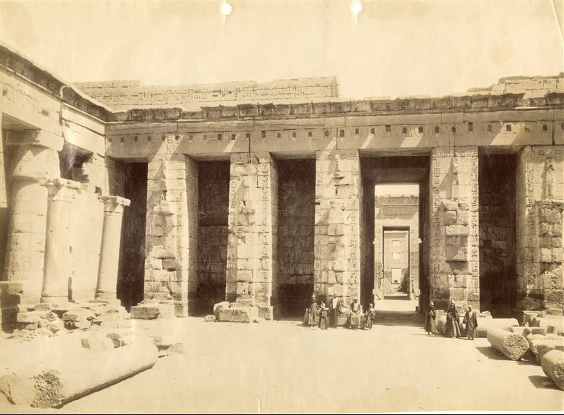 Veduta assiale del tempio di Ramesse III a Medinet Habu, dal secondo cortile in direzione dell’ingresso del tempio. Nel cortile sono ancora prsenti alcune colonne, alcune in piedi, altre a terra. Si intravedono lungo l’asse centrale l’accesso del primo pilone e, in fondo, il migdol. Un gruppo di persone posa per l'autore all'ombra dei pilastri monumentali.  