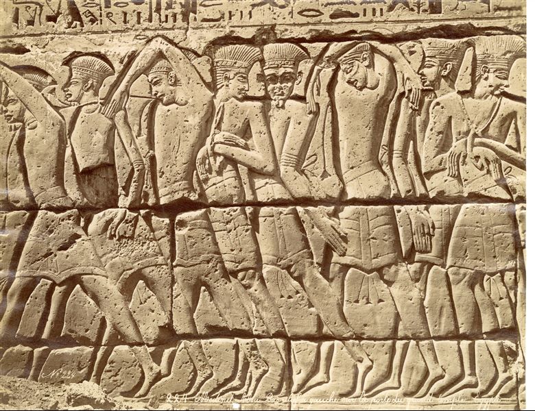 The photograph shows a detail of the decoration of the the first courtyard’s western side in the Temple of Ramesses III at Medinet Habu, West Thebes. It shows a group of prisoners in a row carried by the pharaoh in front of Amun and Mut (who are not visible in this image). 