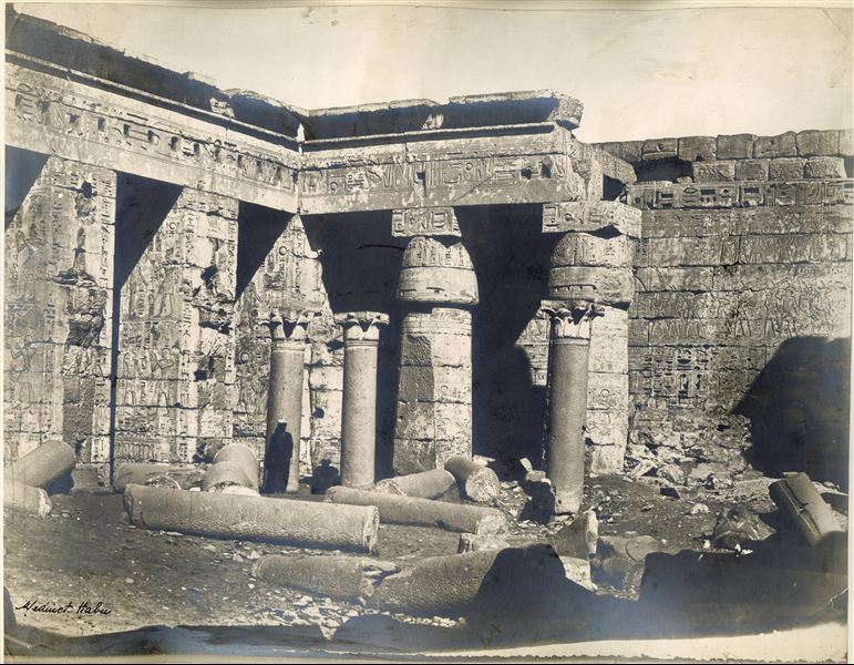 View of the second court of the Temple of Medinet Habu, built by Pharaoh Ramesses III on the west bank of the Nile at Thebes. Several columns can still be seen on the ground, with the standing papyrus columns still partially buried, which help to date the photo to before the site was cleaned and restored. 