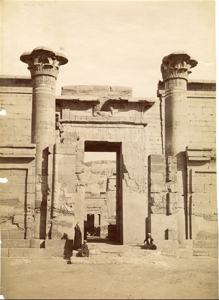 La fotografia mostra tre abitanti del luogo che sostano davanti all’ingresso del cosiddetto Tempio minore, della XVIII dinastia, all’interno del recinto del tempio di Medinet Habu, a Tebe Ovest, costruito dal faraone Ramesse III, della XX dinastia. I portali dell’ingresso, tuttavia, furono costruiti in Epoca tarda e in Età tolemaica. Fotografia scattata dopo la pulizia e restauro del portale. La firma dell'autore è posta in basso a sinistra, a specchio.  