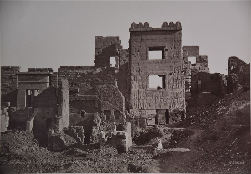 Veduta dell’ingresso monumentale al tempio di Medinet Habu, costruito dal faraone Ramesse III. Il livello del suolo copre ancora parzialmente la parte inferiore dell’antico edificio, chiamato anche migdol (torre fortificata di ispirazione vicino-orientale). Fotografia scattata in direzione dell’esterno, tenendo il I pilone di accesso al tempio alle spalle. La firma dell’autore si trova in basso a destra.  