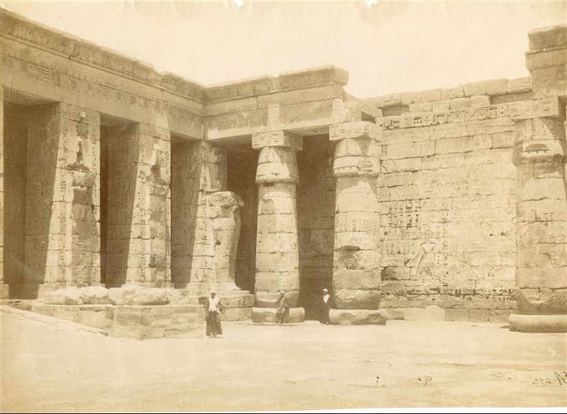 The photograph shows the north-eastern corner of the second courtyard of the Temple of Medinet Habu, with three locals posing for the photographer next to the columns. The document is signed at the bottom right, in mirrored writing but almost illegible. 