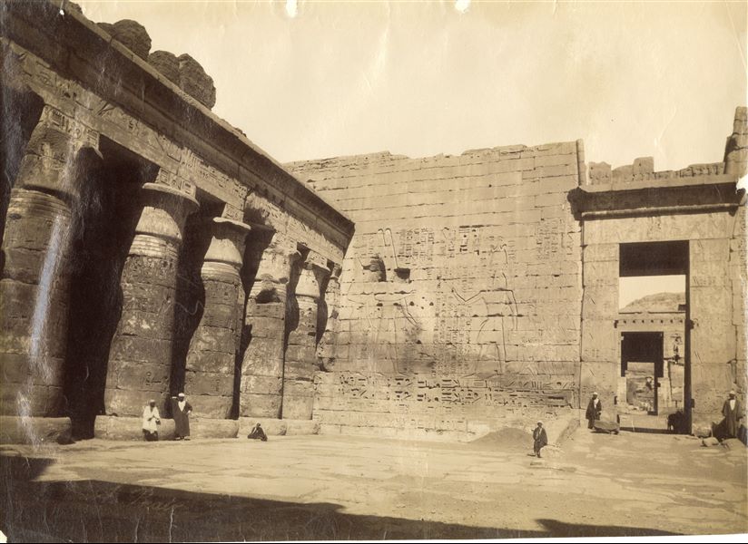 La fotografia, databile a prima del restauro, mostra la facciata (orientale) anteriore del secondo pilone del Tempio di Medinet Habu, costruito dal faraone Ramesse III, decorato con la scena dell'offerta dei prigionieri da parte del sovrano agli dei Amon e Mut. Fotografia scattata dal I pilone. In basso a sinistra, la firma dell’autore.  