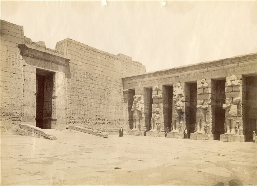 La fotografia mostra l'angolo nord-ovest del primo cortile, con l’ingresso del secondo pilone, del tempio di Medinet Habu a Tebe Ovest, costruito dal faraone Ramesse III. In basso a destra la firma speculare dell’autore.  