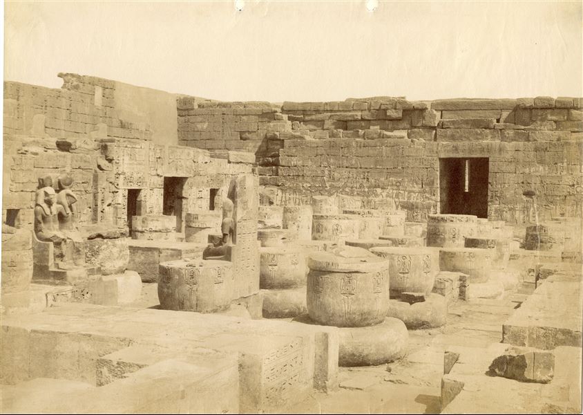 Lo scatto mostra l'angolo sud-occidentale della sala ipostila del tempio di Medinet Habu, costruito dal faraone Ramesse III, a Tebe Ovest. Si riconoscono i resti delle colonne, e, sulla sinistra, due coppie statuarie divine (di una si vede solo retro e il profilo di una divinità), gli ambienti laterali e la porta assiale. Il pilone si intravede oltre la parete di fondo. Sulla base delle annotazioni sul retro è possibile attribuire lo scatto ad Antonio Beato.  