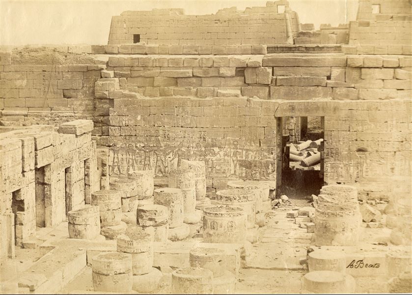 The photograph shows the south-western corner of the hypostyle hall from the Temple of Medinet Habu, built by Pharaoh Ramesses III in west Thebes. In the foreground, the remains of the columns and the side rooms. In the background, the top of the entrance pylon to the complex, and the columns that are still on the ground. The author's signature is at the bottom right. 