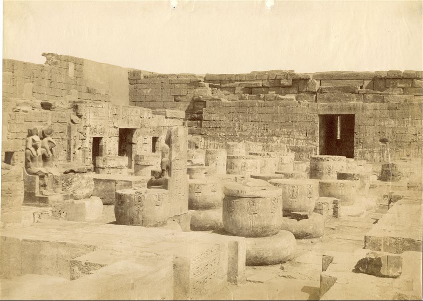The photograph shows the south-western corner of the hypostyle hall of the Temple of Medinet Habu, built by Pharaoh Ramesses III, in west Thebes. The remains of the columns are visible and on the left, two pairs of divine statues (of one, only the back and profile of the deity can be seen), the side rooms and the axial door. The pylon can be glimpsed beyond the back wall. Based on the annotations on the back, the image can be attributed to Antonio Beato. 