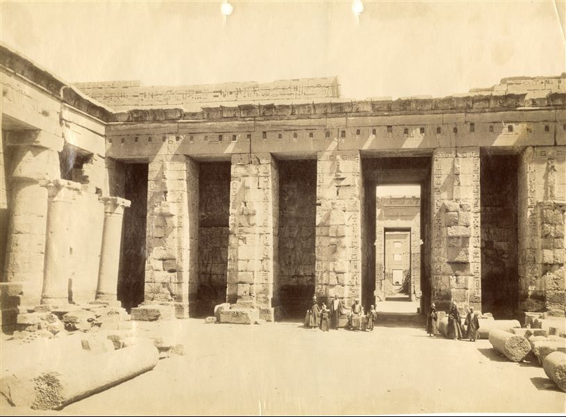 Axial view of the Temple of Ramesses III at Medinet Habu, from the second courtyard in the direction of the temple entrance. A number of columns are still present in the courtyard, some standing, others on the ground. Along the central axis is the entrance to the first pillar and the migdol a the back. A group of people pose for the photographer in the shadow of the monumental pillars. 