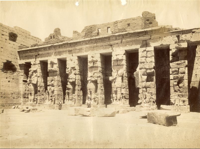The photograph depicts the north-eastern side of the first courtyard of the temple complex of Medinet Habu in West Thebes, built by Ramesses III. Here the statues leaning against the pillars of Pharaoh Ramesses III stand out. Two locals position themselves to show the colossal size of the statues. Towards the left side,  the photograph is slightly blurred. 