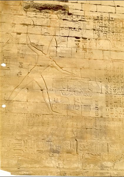 The photograph shows a detail of the decoration of the left side of the entrance pylon of the Temple of Medinet Habu, built by Pharaoh Ramesses III. The ruler wields a mace to strike down enemies whilst holding them by the hair with his left hand. This is a typical decorative theme portryed on temple exteriors. The author's signature can be found in the bottom right, in a mirror-like handwriting, and is almost illegible. 