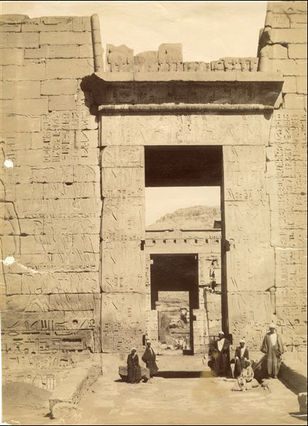 Veduta in primo piano dell'accesso al secondo cortile del tempio funerario di Medinet Habu, costruito dal faraone Ramesse III, con un gruppo di abitanti locali in posa per l'autore. La firma dell’autore è in basso a sinistra. 