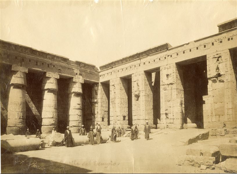 La fotografia riprende l'angolo sud-orientale del secondo cortile del tempio di Medinet Habu, costruito dal faraone Ramesse III, con alcuni abitanti locali che guardano verso l’obiettivo. La corte presenta ancora diverse colonne a terra. Questo rende lo scatto antecedente alla fotografia Inv11_004. La firma dell'autore è apposta in basso a sinistra.  