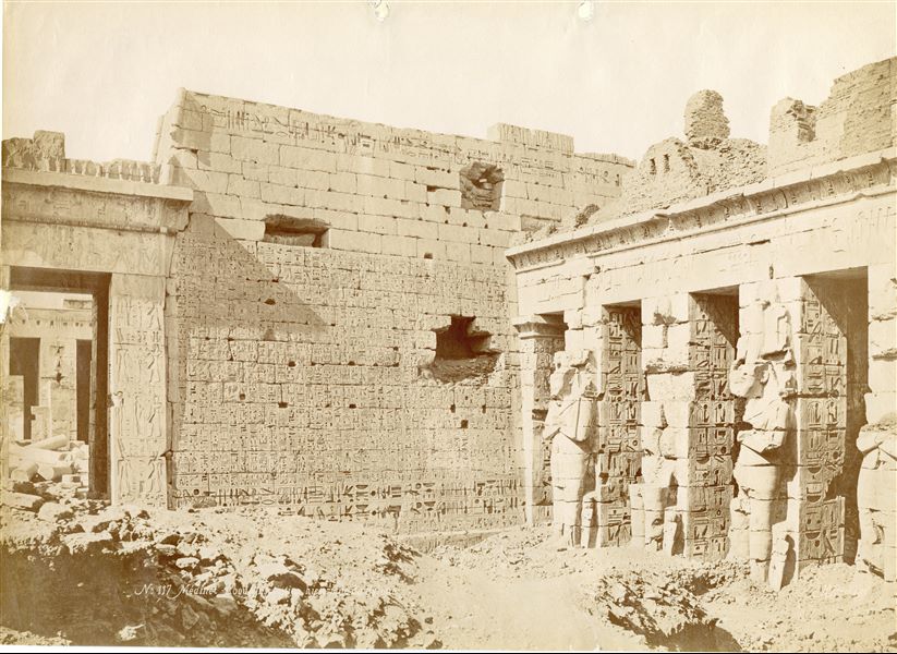 The photograph shows the east façade (north side) of the second pylon of the Temple of Medinet Habu in West Thebes, built by Ramesses III, and the first courtyard of the temple. Note that the original ground level is still covered by sand and debris, which cover the bases of the statues and form an uneven surface in the courtyard. The author's signature is at the bottom right. 