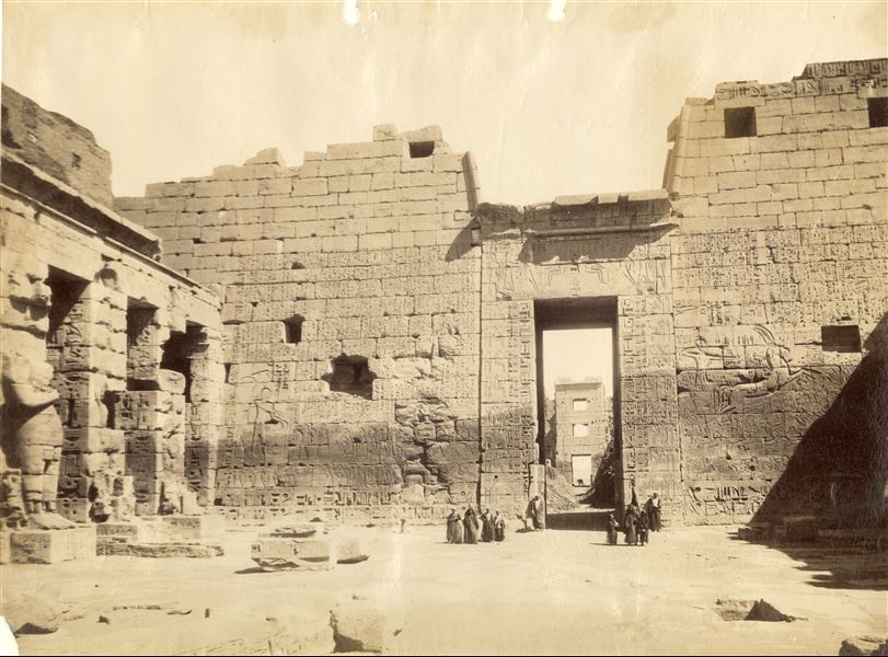 The photograph shows the rear (western) façade of the first pylon of The temple of Medinet Habu in West Thebes photographed from the first courtyard, with locals nearby. Through the gateway, the watchtower is visible, a structure named after the syrian fortresses of the time called a migdol. The author's signature is almost illegible, but present at the bottom right-hand corner. 