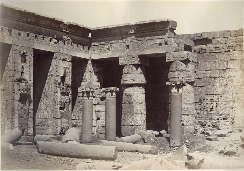 Veduta della seconda corte del tempio di Medinet Habu, costruito dal faraone Ramesse III sulla riva ovest del Nilo, a Tebe. Si notano ancora numerose colonne a terra, indizio che la fotografia è stata scattata prima dei lavori di pulizia e restauro del sito. In basso, la firma dell’autore.  