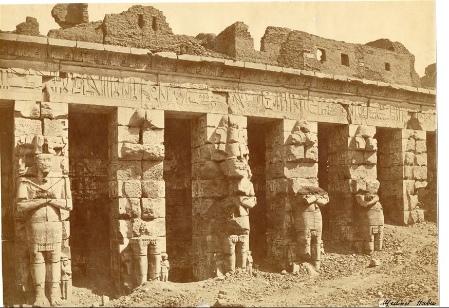 The image shows a view of the northern colonnade in the first courtyard of Medinet Habu, with the Osiride pillars leaning against some columns, not yet fully cleared of sand and debris. 