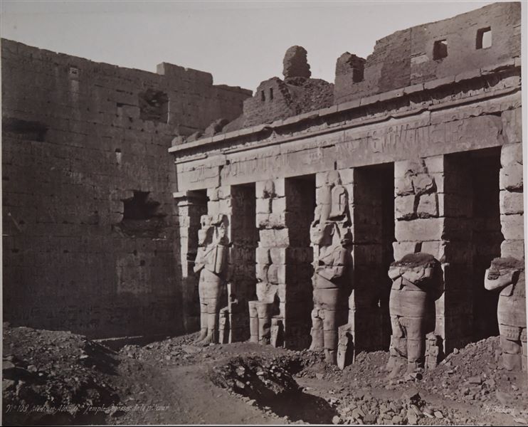Veduta della prima corte del tempio di Medinet Habu, costruito dal faraone Ramesse III sulla riva ovest del Nilo, a Tebe. I pilastri osiriaci ancora parzialmente interrate dimostrano che la data dello scatto è anteriore ai lavori di pulizia e restauro del sito. In basso a destra, la firma dell’autore.  